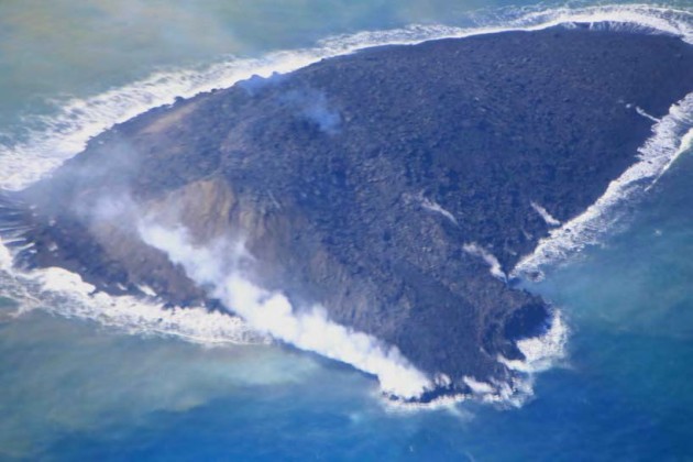 写真２。新島の全景と溶岩流。2013 年11 月24 日昼前。 黒色の溶岩流が中腹から流れ手前の海に流入。右（北）側に古い流れでできたと思われる土手が発達。左手前（南東）と左奥（南西）に斜 面崩壊でできた褐色の崖が発達している。青白い噴煙が頂部火口から上がっている。東から撮影。
